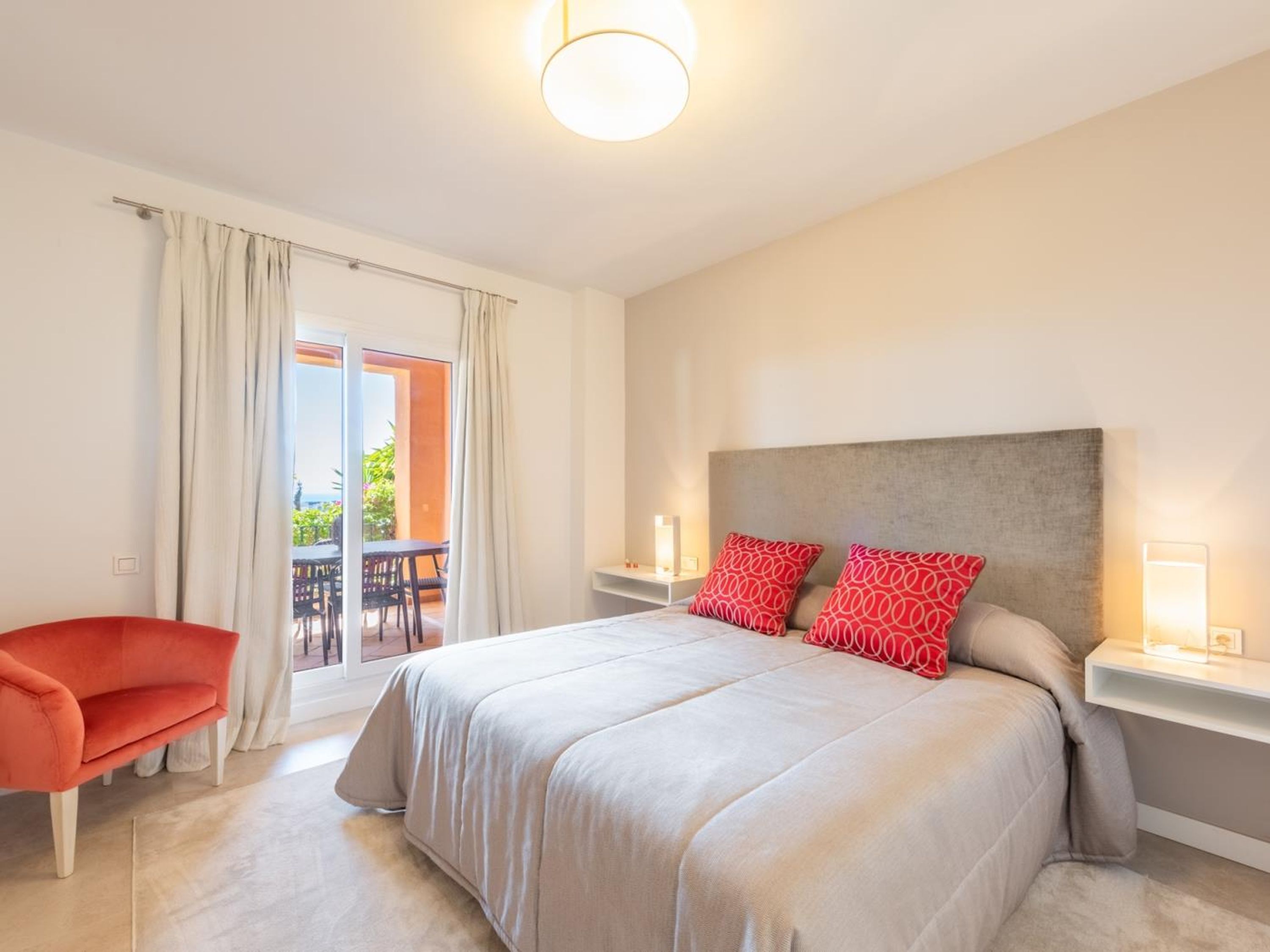 Quarto moderno com uma cama grande, uma cadeira vermelha, varanda com assento externo e vista para área verde.