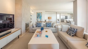 A modern living room with a flat-screen TV, a white coffee table with magazines, and a beige sofa.
