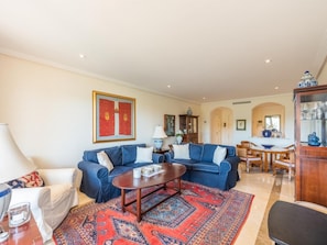 A living room with two blue sofas, a coffee table, a patterned rug, and a dining area with a table and chairs.