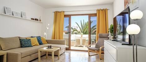 A modern living room with a sofa, coffee table, and armchair, a flat-screen TV, and a balcony with yellow curtains.