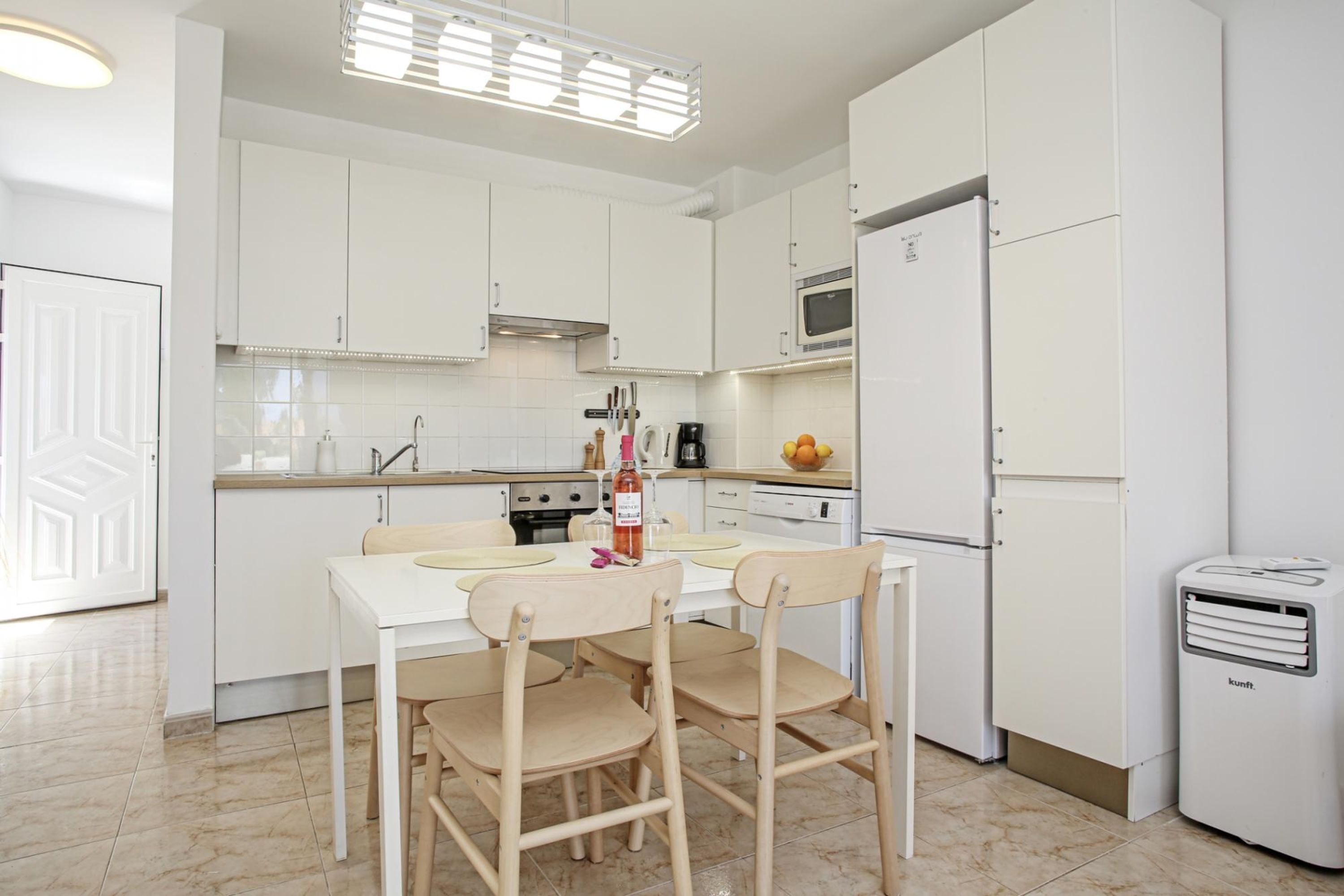 A modern kitchen with white cabinets, a dining table with chairs, a microwave, a refrigerator, and a trash bin.