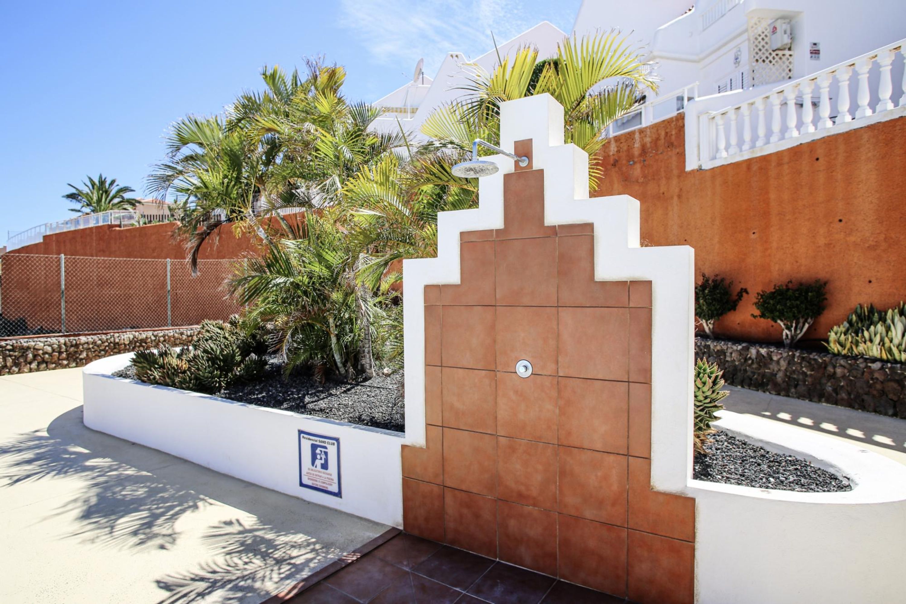 A gated entrance with a white and brown wall, palm trees, and a clear sky.