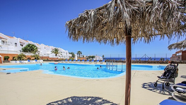 A pool area with lounge chairs, a thatched umbrella, and a view of the sea. - Sand Club Apartment 202 (San Miguel de Abona)