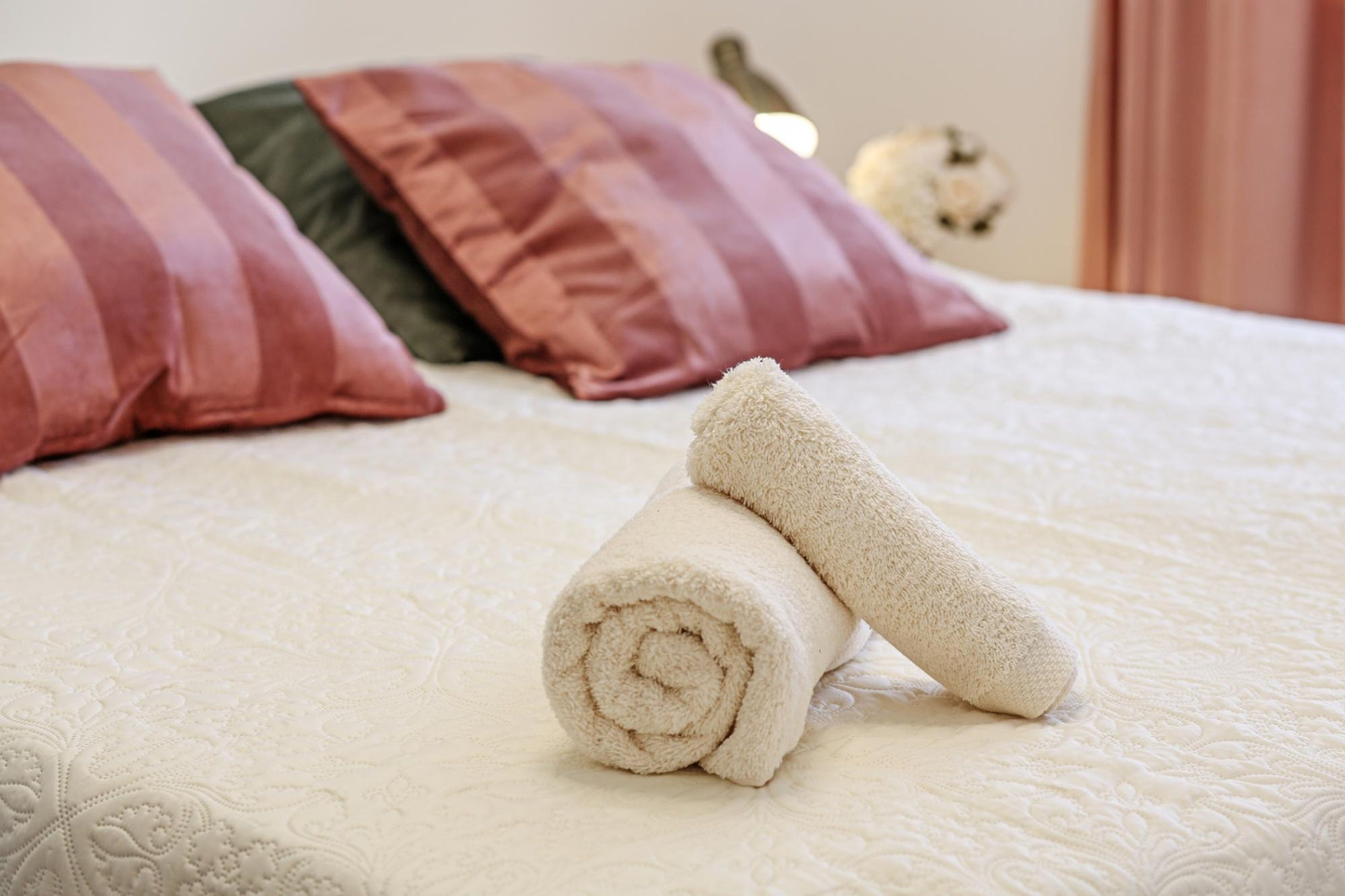 A neatly made bed with a rolled white towel and a folded white towel on a textured white bedspread.