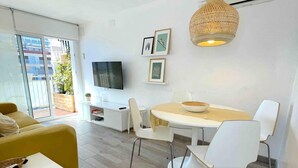 A modern living room with a sofa, a dining table with chairs, a TV mounted on the wall, and a large window with a view of buildings and greenery. - Fabuloso Apartamento Cerca Playa y Portaventura (Salou)
