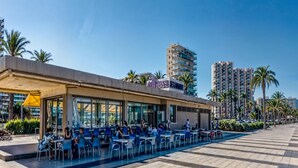 An outdoor cafe with tables and chairs, a building with a sign, palm trees, and high-rise buildings in the background. - Apartamento N Utico (Alicante)