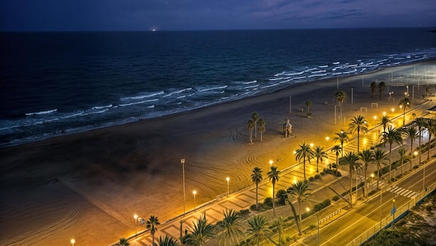 A beach with a paved walkway, palm trees, and a distant view of the ocean at night. - Apartamento N Utico (Alicante)
