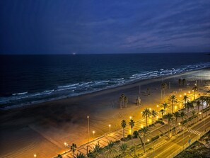 A beach with a paved walkway, palm trees, and a distant view of the ocean at night. - Apartamento N Utico (Alicante)