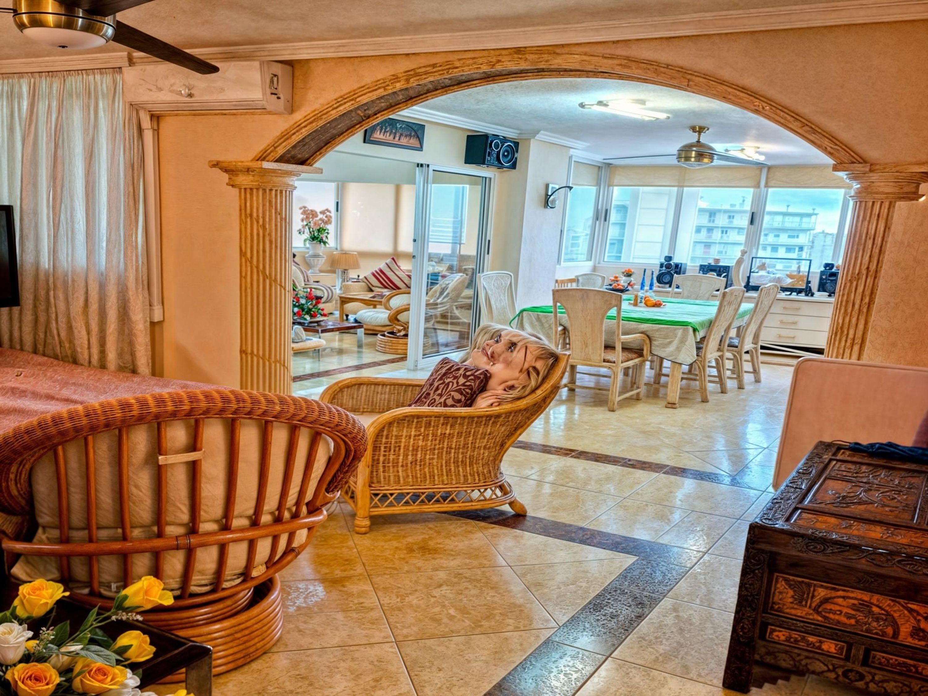 A room with a wicker chair, a wooden chest, and a dining area with a green tablecloth.