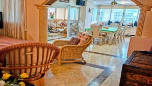A room with a wicker chair, a wooden chest, and a dining area with a green tablecloth. - Apartamento N Utico (Alicante)