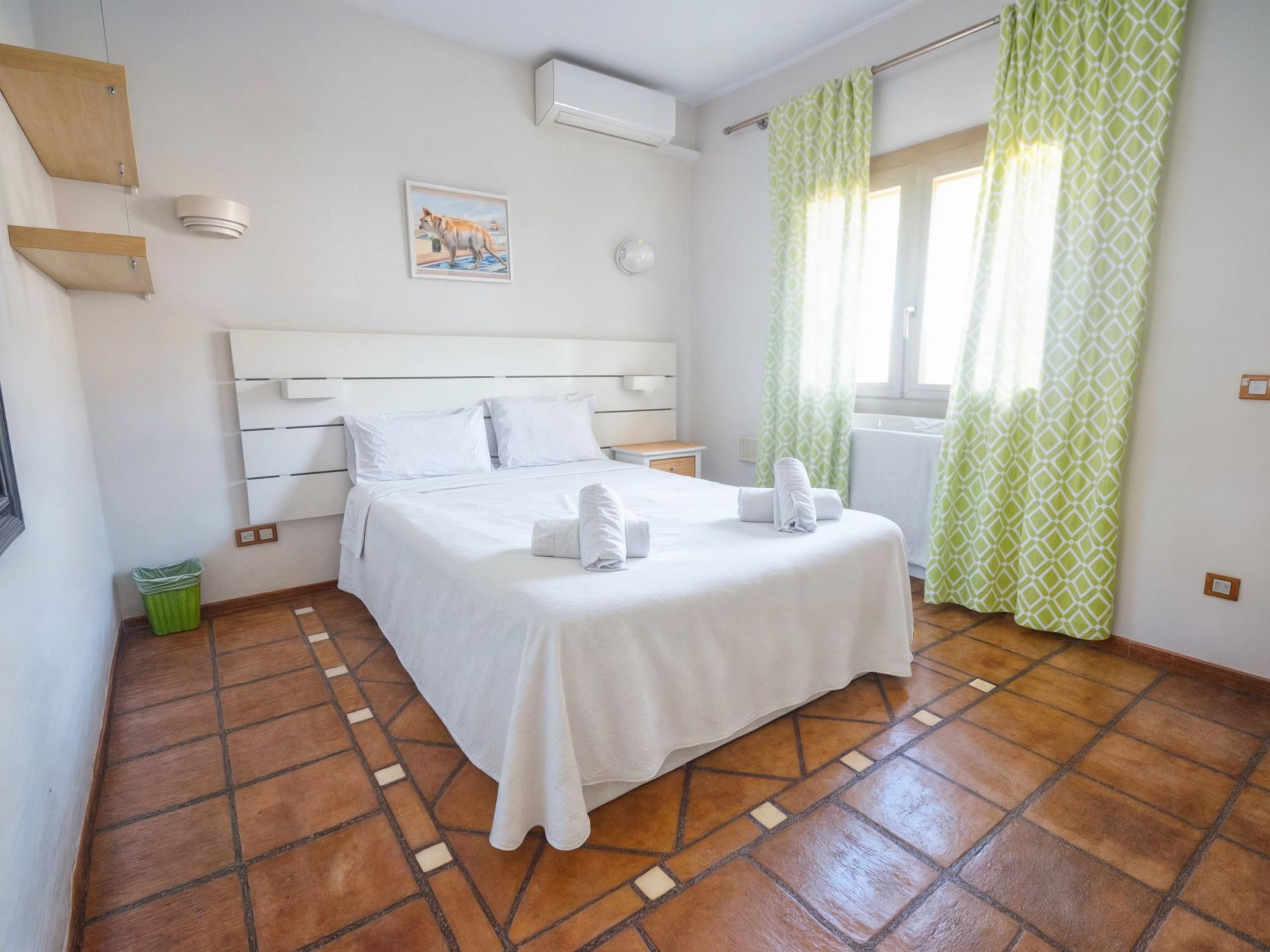 A neatly arranged bedroom with a bed, a small bedside table, a wall-mounted air conditioner, and a window with patterned curtains.