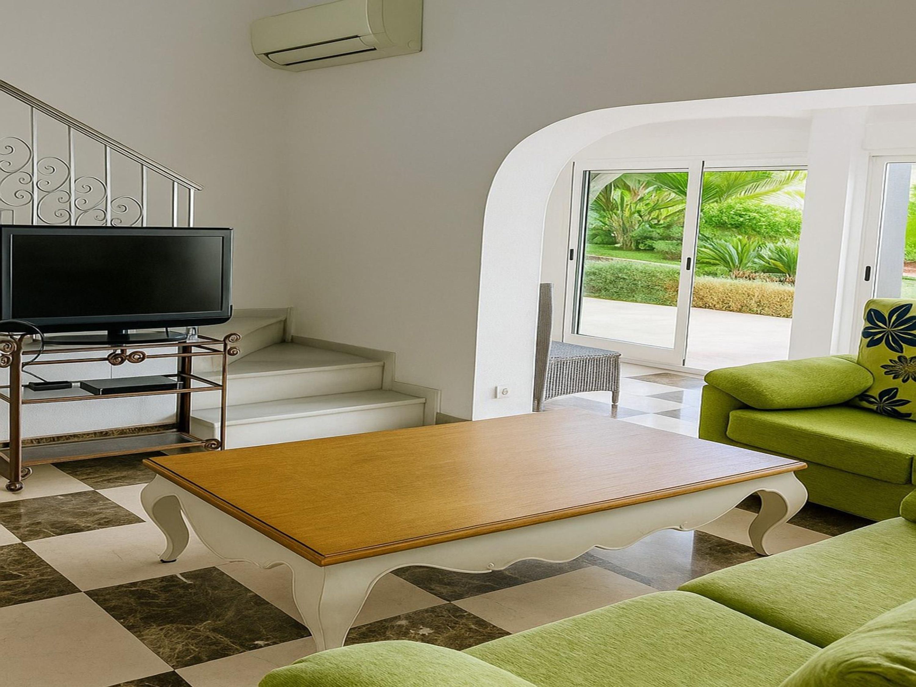 Un salon équipé d’un téléviseur à écran plat, d’une table basse en bois, d’un canapé vert et d’un escalier menant à un balcon offrant une vue sur la verdure.