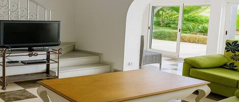 Un salon équipé d’un téléviseur à écran plat, d’une table basse en bois, d’un canapé vert et d’un escalier menant à un balcon offrant une vue sur la verdure.