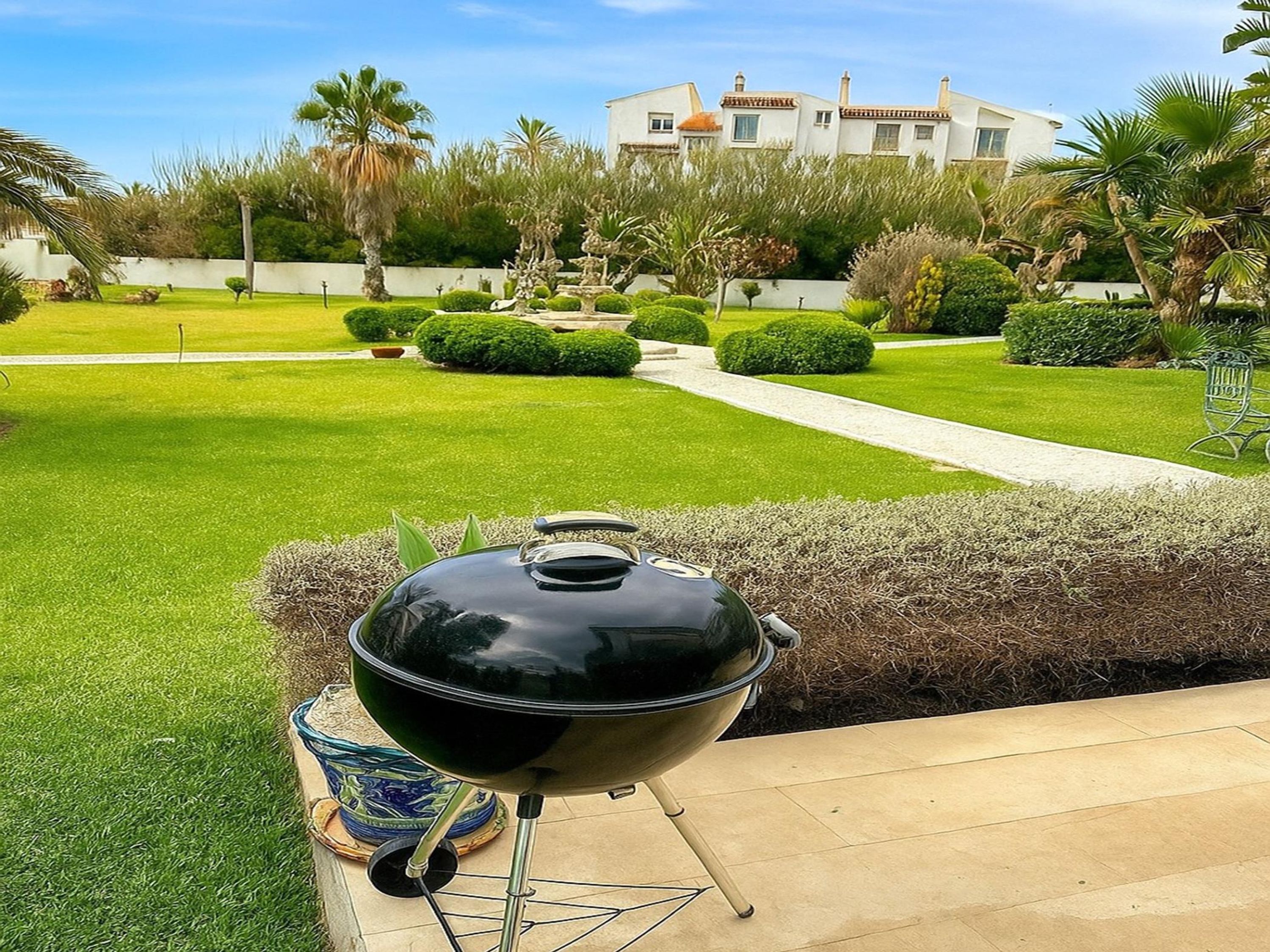 A barbecue grill in a well-maintained garden with a pathway, neatly trimmed hedges, and a building in the background.