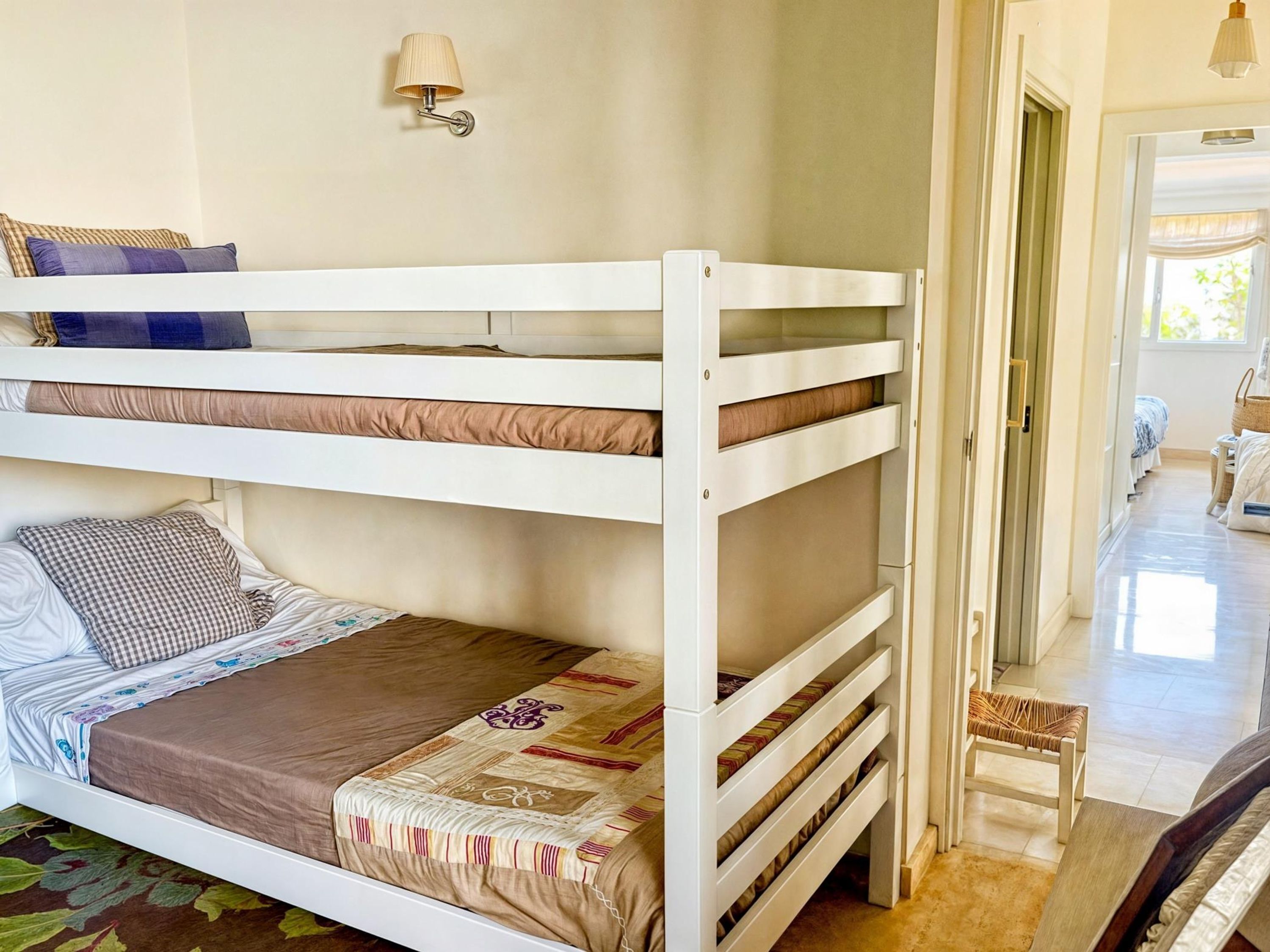 A bunk bed room with a white color scheme, a wall-mounted lamp, and a view of the hallway.