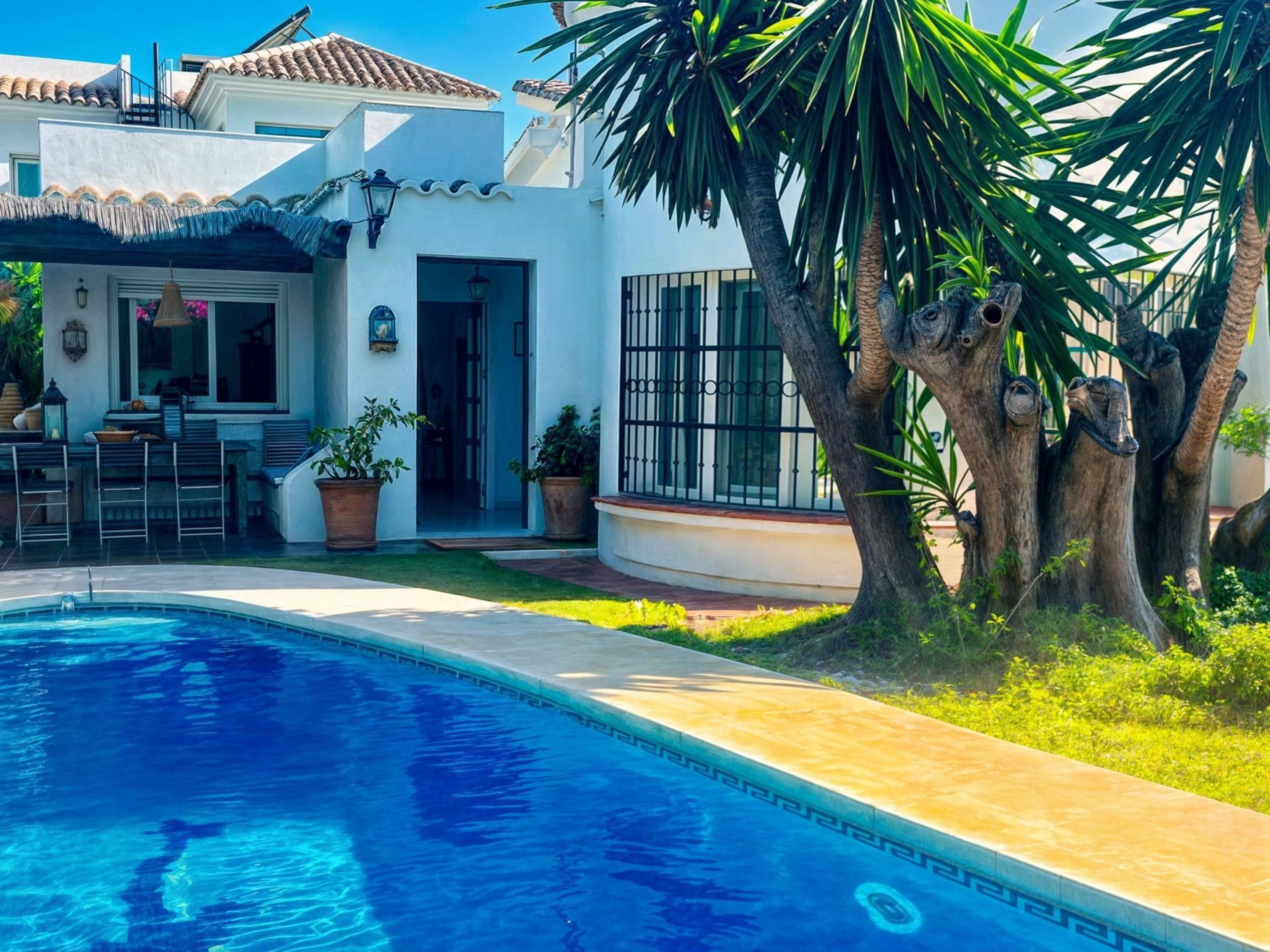 A pool area with a clear blue pool, a patio with outdoor furniture, and a white building with a tiled roof.