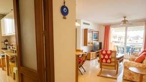 A cozy living room with a wooden door, a dining area with a table and chairs, a TV, and a balcony with blue chairs. - Apartamento Parque Central (Estepona)