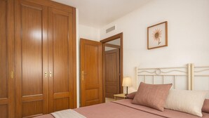 A bedroom with a large bed, wooden wardrobe, and a nightstand with a lamp. - Apartamento Parque Central (Estepona)