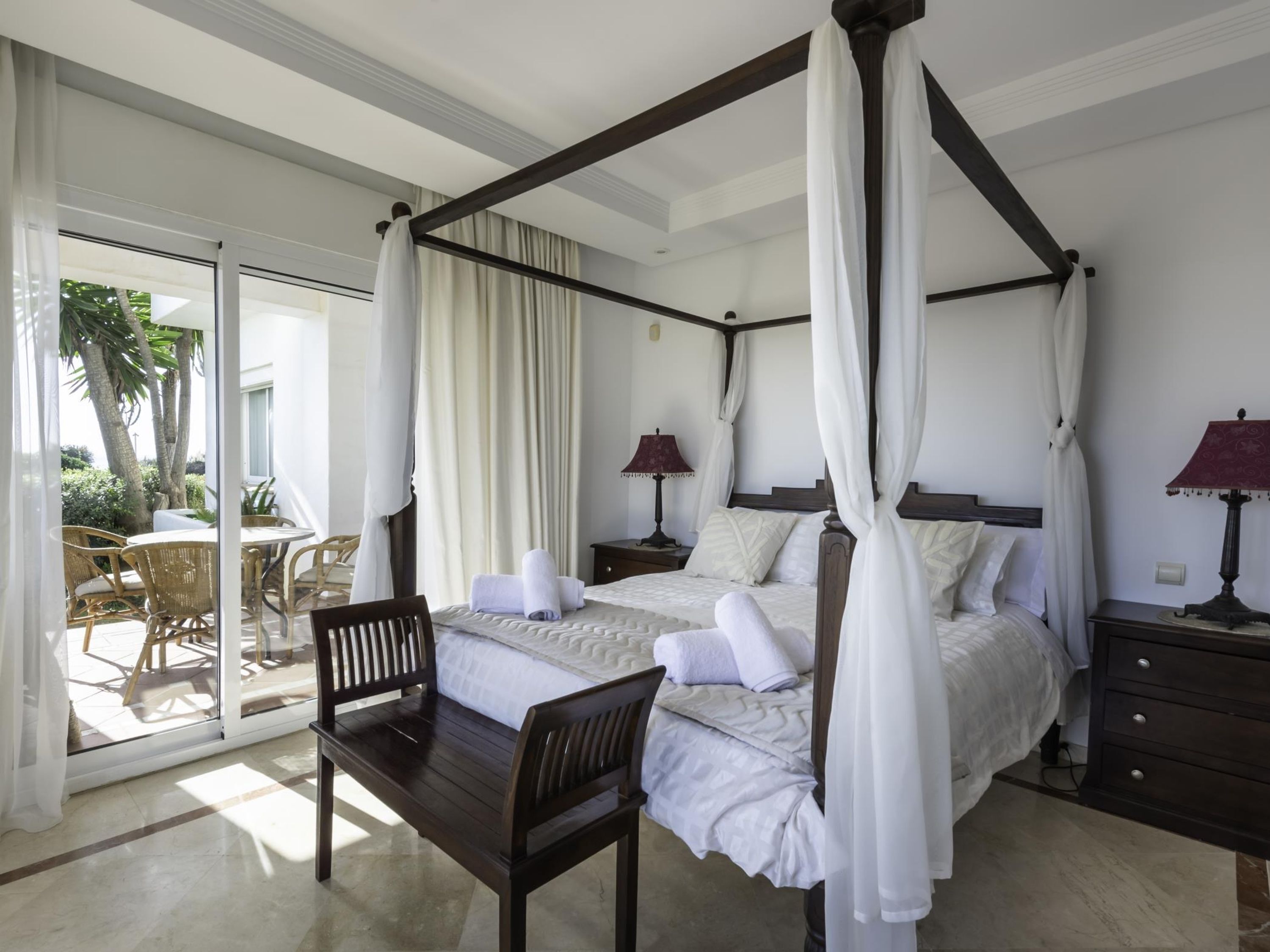 A four-poster canopy bed with white linens, a wooden nightstand with a lamp, and a dining area with wicker chairs and a table.