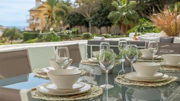 A table set for dining with white bowls, plates, and wine glasses, surrounded by greenery and a view of a residential area.