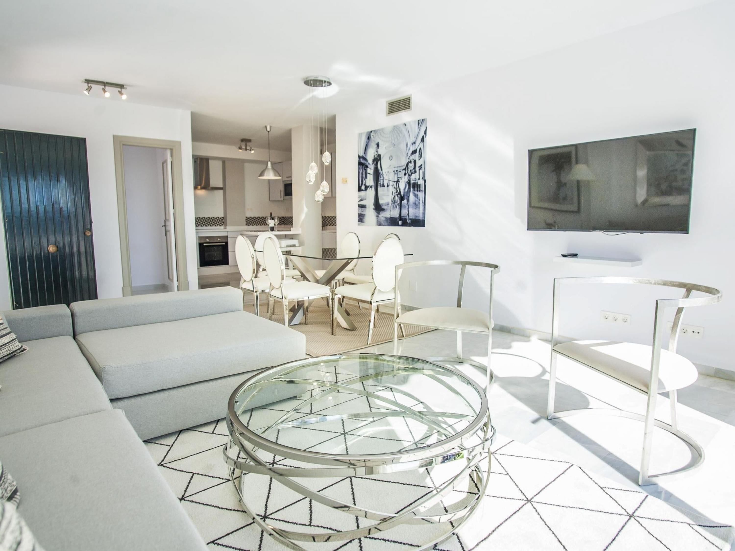 Una sala de estar moderna con una mesa de centro de vidrio, un sofá y una zona de comedor con sillas.