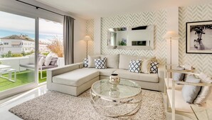 A modern living room with a glass coffee table, a sofa with patterned cushions, a large mirror, and a floor lamp.