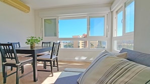 Una habitación luminosa con una mesa de comedor, sillas y un sofá. Hay ventanas amplias que dan a vistas de edificios.