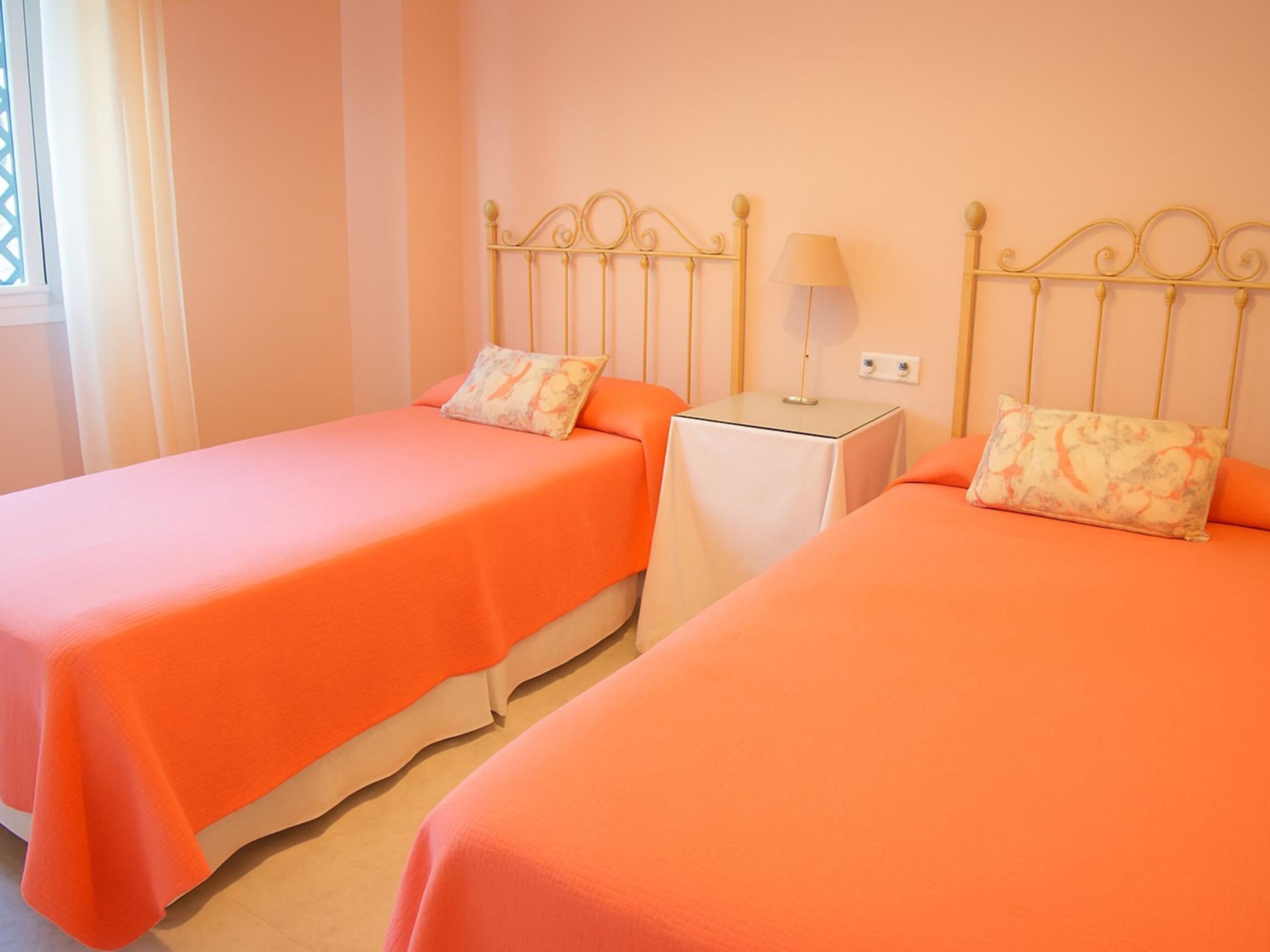 Two twin beds with matching orange bedding and floral-patterned pillows, a small bedside table with a lamp, and a window with curtains.