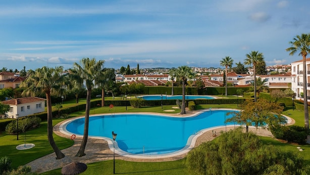 A large, curved swimming pool surrounded by well-maintained lawns and palm trees, with a view of residential buildings in the background.