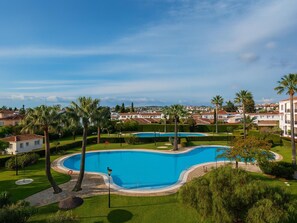 Uma grande piscina curvada, rodeada por relvados bem cuidados e palmeiras, com vista para edifícios residenciais ao fundo.