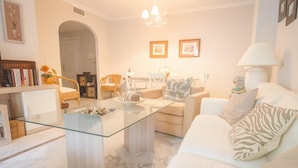 A modern living room with a glass coffee table, white sofas, a bookshelf, and a dining area.