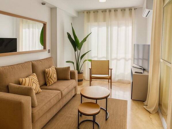 A modern living room with a sofa, two chairs, a small table, a large plant, a TV, and a window with curtains. - Apartamento Maite in Estepona (Estepona)