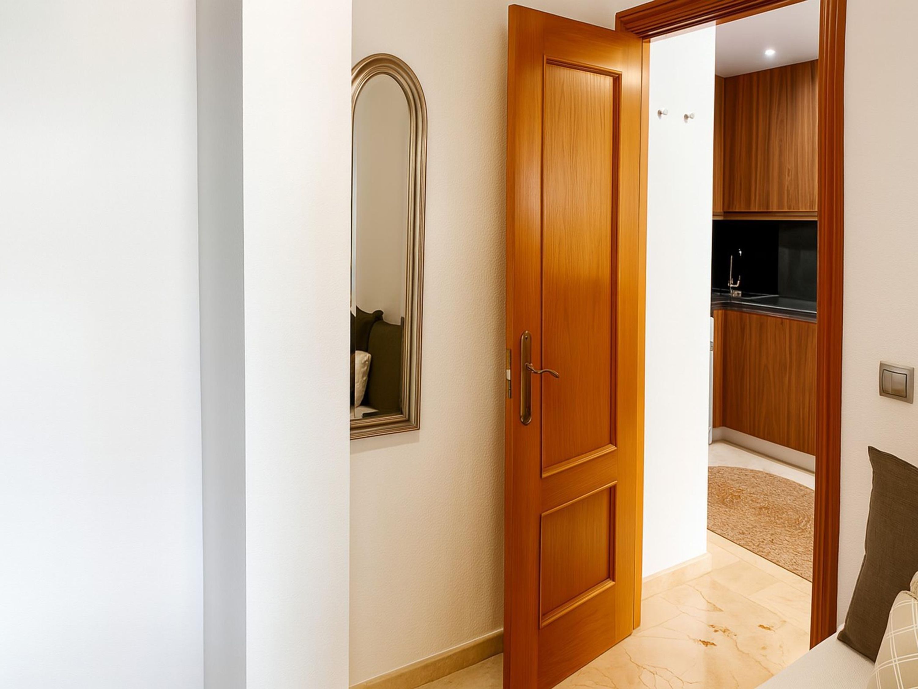 Une porte en bois avec un miroir, qui donne sur un espace cuisine aménagé de meubles en bois et doté d’un plan de travail noir.