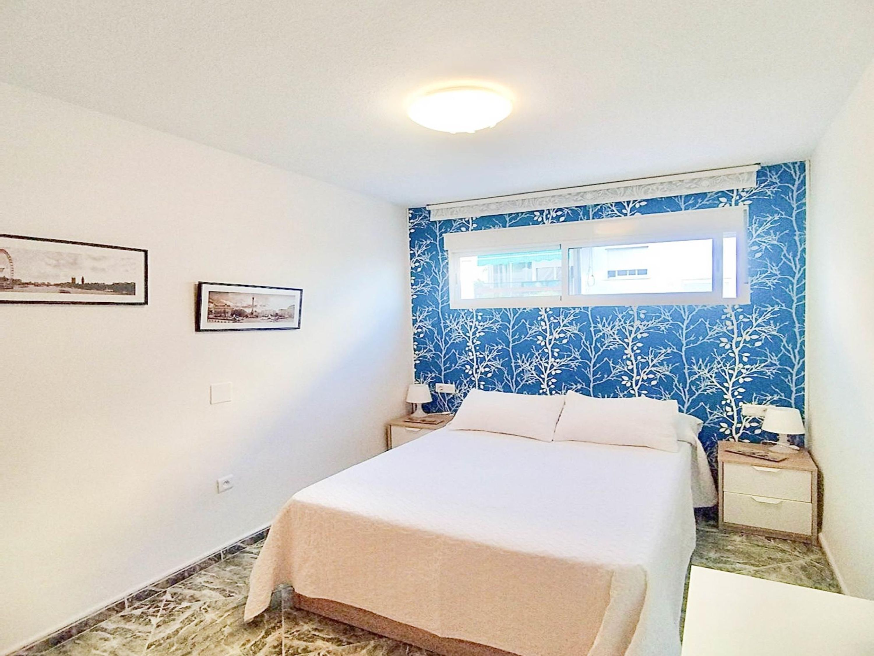 Une chambre à coucher avec un lit, deux tables de chevet, une fenêtre et deux tableaux encadrés accrochés au mur.