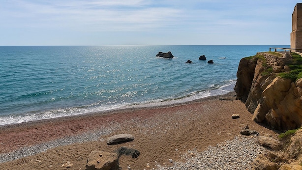 Ein Küstenstrand mit einer felsigen Klippe und einem Gebäude auf der rechten Seite.