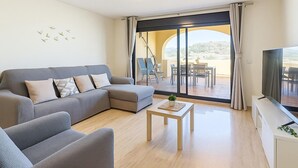 Ein modernes Wohnzimmer mit einer grauen Eckcouch, einem kleinen Holztisch mit einer Pflanze und einem Fernseher auf einer weißen Kommode. Es gibt einen Balkon mit Tisch und Stühlen und einen Blick ins Freie.