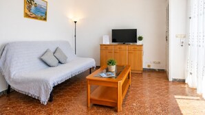 A living room with a sofa, a coffee table, a television on a wooden cabinet, a ceiling fan, and a painting on the wall. - Apartamento Acogedor en Primera Linea Liliane (Sitges)