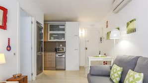 Een knus woongedeelte met een kitchenette, een grijze bank met groene, patroonrijke kussens en een kleine eettafel met een groene placemat.