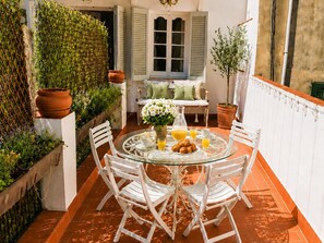 Una mesa con superficie de vidrio y sillas blancas, una mesita redonda con mantel blanco y una ventana con contraventanas.