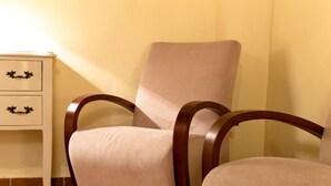 Uma cadeira com assento acolchoado e apoio de braço de madeira, posicionada ao lado de uma mesa de cabeceira branca com duas gavetas.