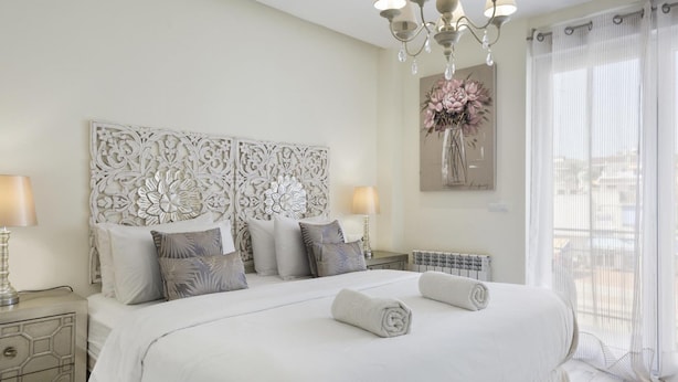 Ruime slaapkamer met een groot bed, decoratief hoofdeinde, nachtlampjes en uitzicht op het groen door dunne gordijnen.