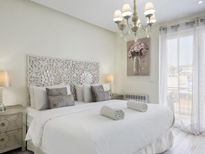 A spacious bedroom with a large bed, decorative headboard, bedside lamps, and a view of the outdoors through sheer curtains.