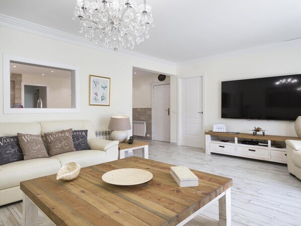 A modern living room with a white sofa, a wooden coffee table, a chandelier, a flat-screen TV, and a fireplace.