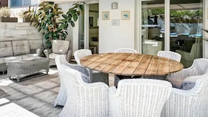 Een terras met een ronde houten tafel, omringd door rieten stoelen, een stenen muur en potplanten.