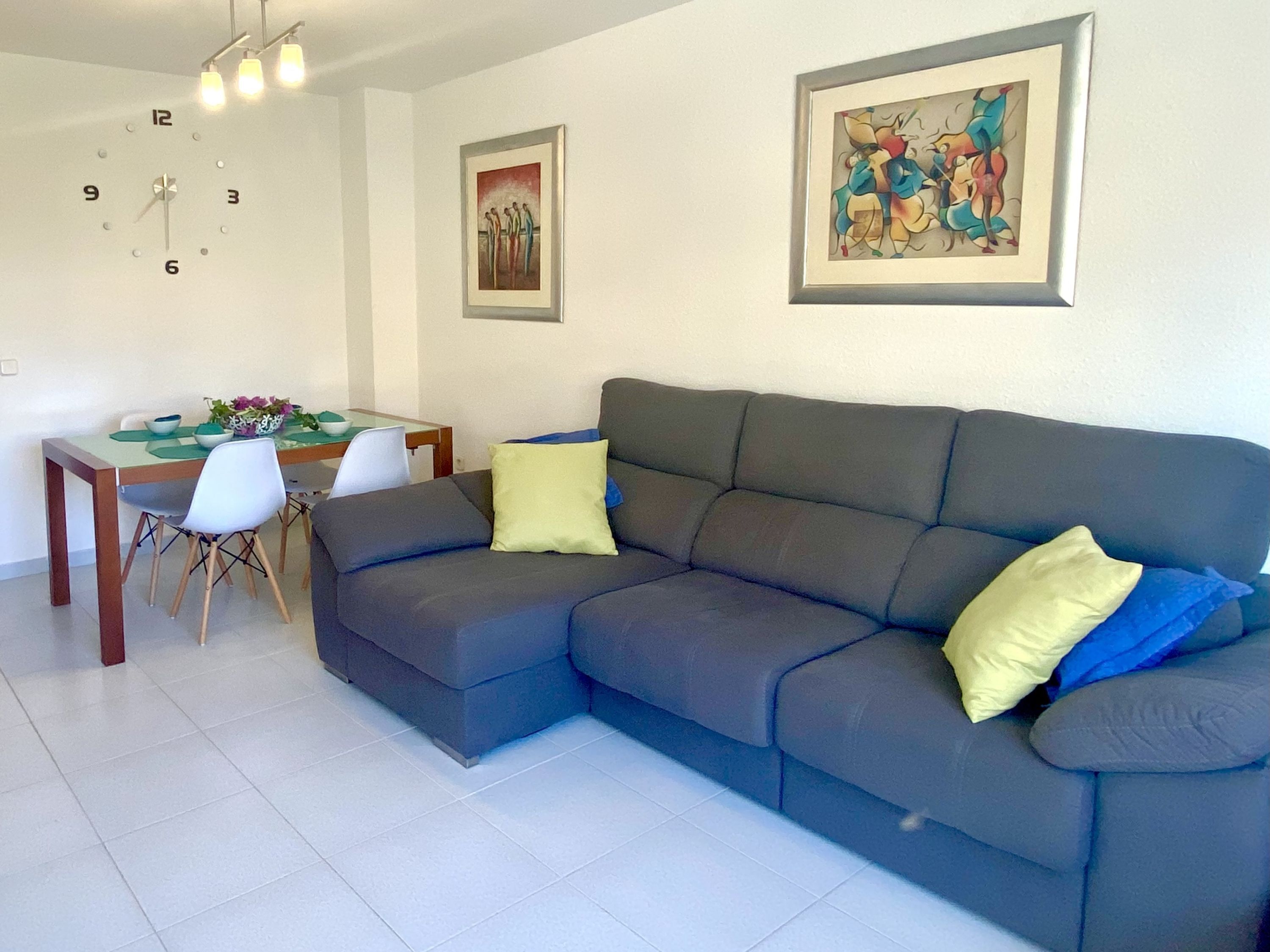 A modern living room with a grey sofa, a dining table with chairs, and a wall clock.