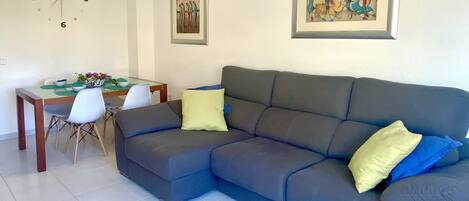 A modern living room with a grey sofa, a dining table with chairs, and a wall clock.