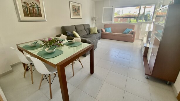 A modern living room with a dining area, featuring a sofa, a table set with a vase of flowers, and artwork on the walls. - Rental Unit in Calafell 3 Bedrooms 3 Beds 1 (El Vendrell)