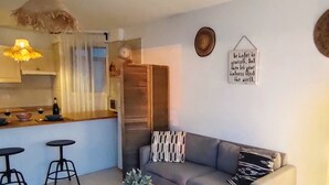 Un salon comprenant un canapé, deux tabourets de bar, une petite table avec une plante, un coin cuisine avec un comptoir et une horloge, ainsi qu’une citation inspirante sur le mur.