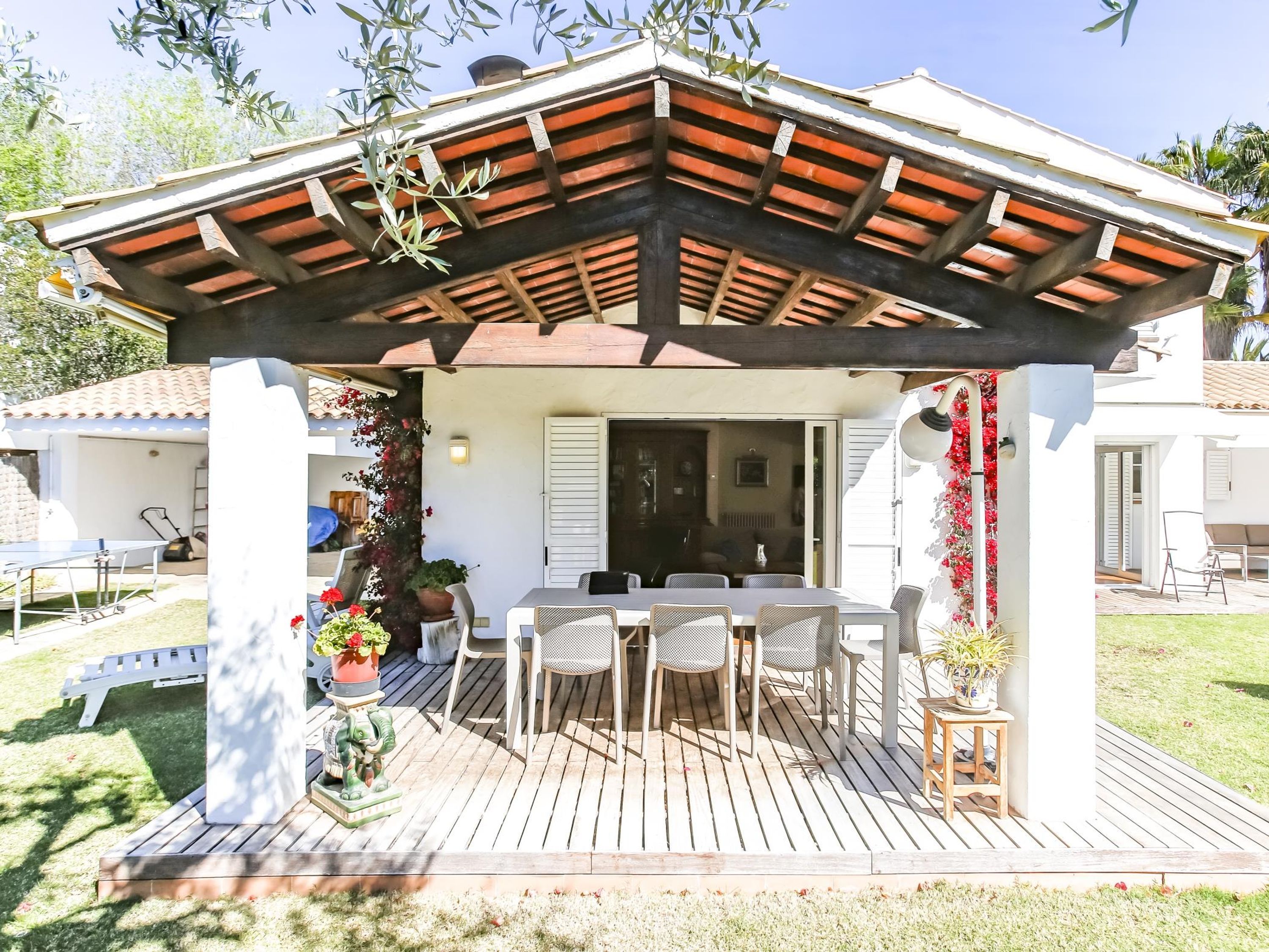 Eine überdachte Terrasse im Freien mit Holzboden, weißen Wänden und einer Pergola mit Ziegeldach.
