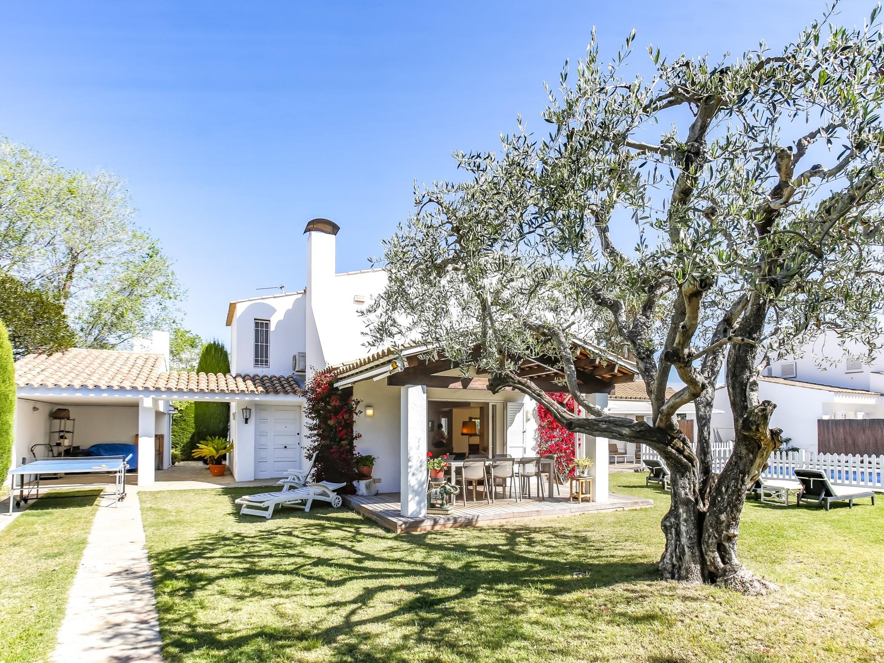 A white villa with a tiled roof, a covered patio with outdoor furniture, and a large tree in the foreground.